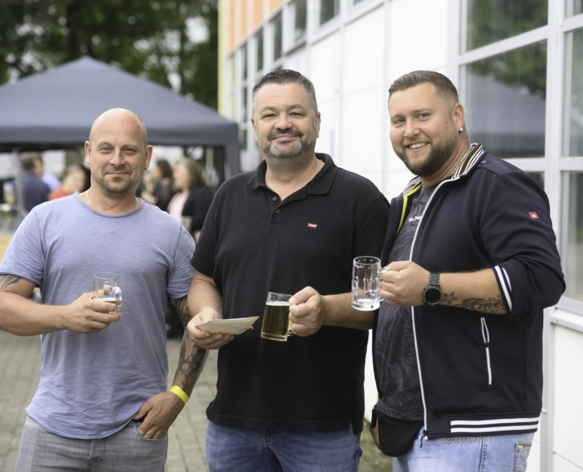 3 Männer blicken auf dem Sommerfest gut gelaunt in die Kamera und halten jeweils ein Glas Bier in der Hand