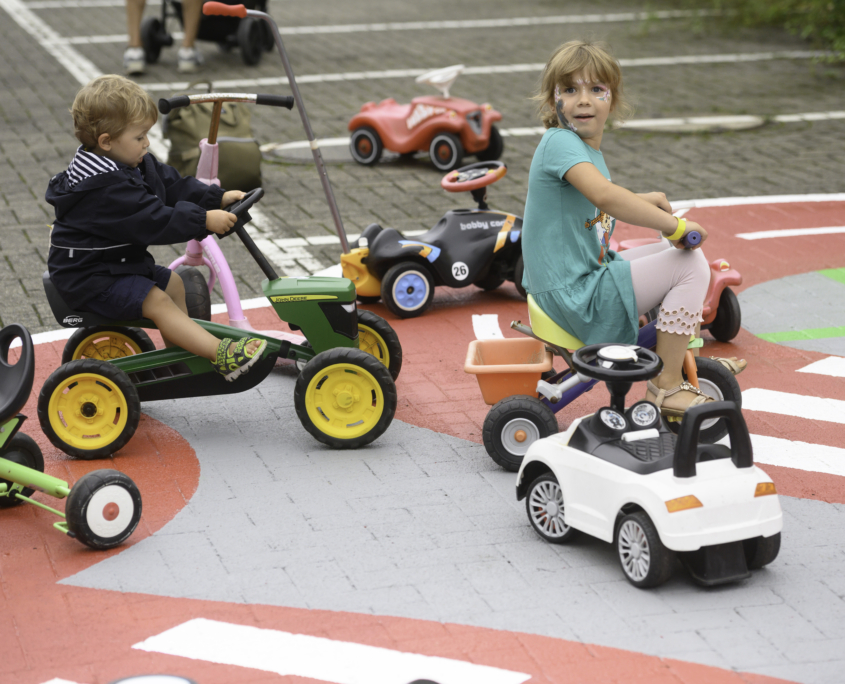 Zwei Kinder fahren auf einem Sommerfest auf Bobby Cars auf einer dafür vorgesehen bunten Farbahnmarkierung