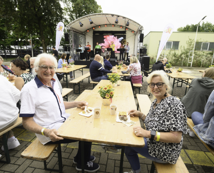 Zwei Senioren blicken auf einem Sommerfest auf einer Bierzeltgarnitur sitzend in die Kamera. Im Hintergrund spielt eine Brassband auf einer Bühne.