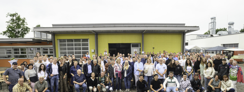 Gruppenbild der Neustädter Belegschaft auf dem Sommerfest vor dem alten Lager und einer gelben Wand